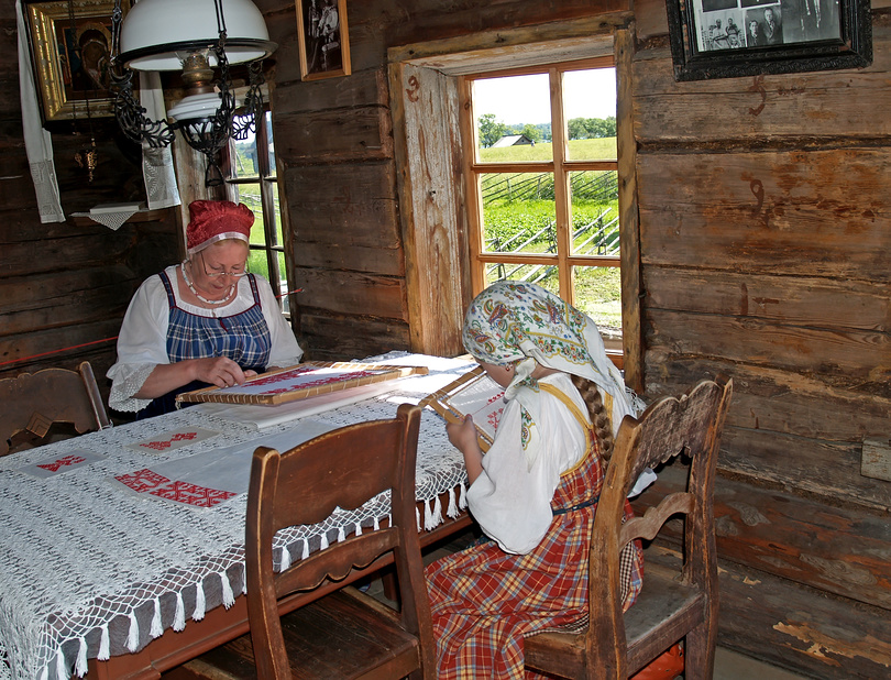 Две женщины в традиционной одежде вышивают за деревянным столом в деревенской избе.