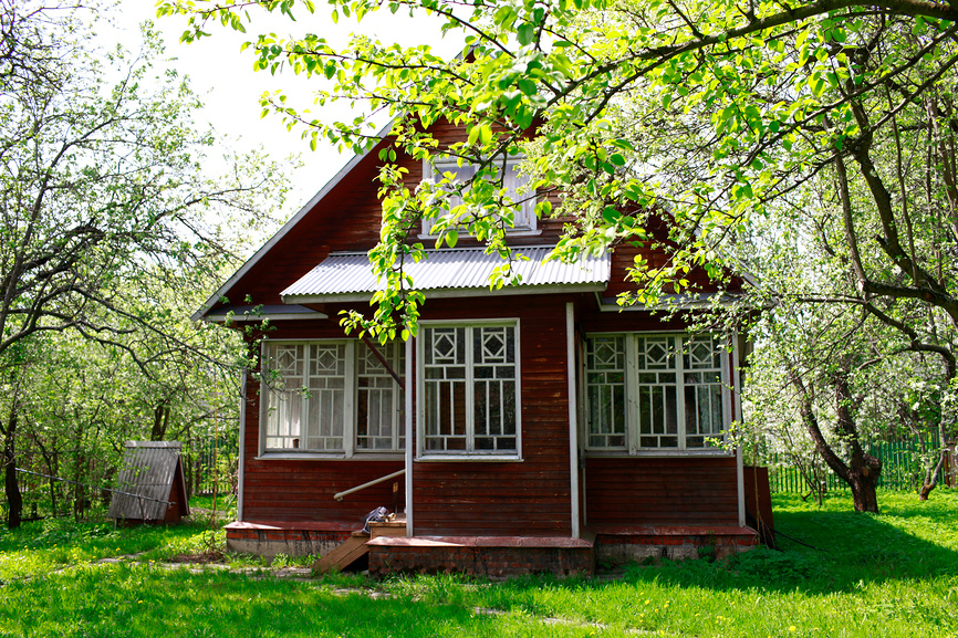 Деревянный дачный домик, красная обшивка, резные окна, зелёная листва, солнечная поляна.