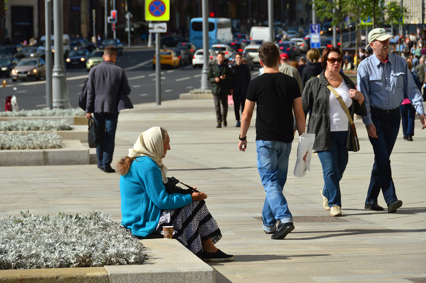 На переднем плане изображении сидит пожилая женщина, городская улица с оживлённым движением пешеходов и транспорта