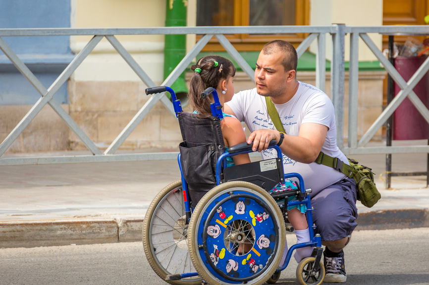 Больной ребенок-инвалид в инвалидной коляске в окружении отца на городской улице.